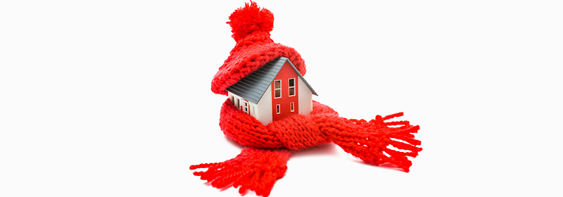 A model of a home, with a red knitted hat on its roof, and a red knitted scarf wrapped around it, on a white background.