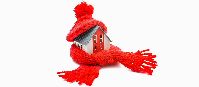 A model of a home, with a red knitted hat on its roof, and a red knitted scarf wrapped around it, on a white background.