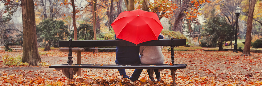 A couple sitting on a park bench in Autumn. They are facing away from the viewer, and are sharing a bright red umbrella.